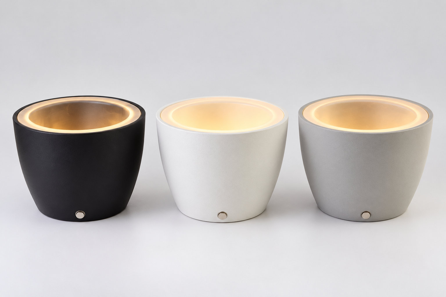 Three actual LumaHalo planter prototypes in Charcoal Gray, Warm White and Mist Gray, with illuminated upper interiors and small circular components near the lower front.